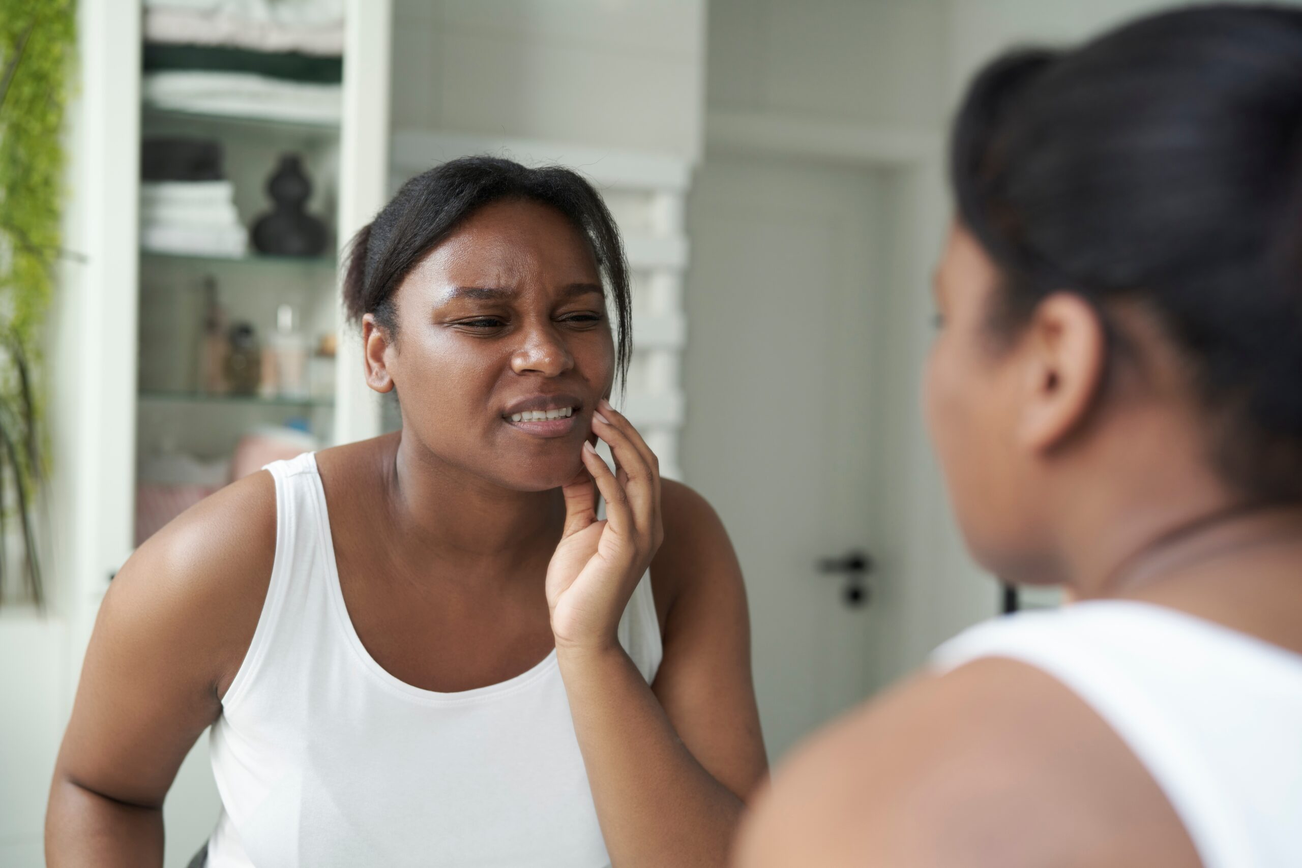 young woman looking in the mirror holding her jaw in pain, bruxism signs