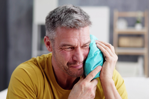 man in a yellow shirt holding a blue ice pack on his jaw, severe tooth pain